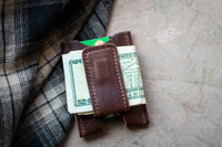 Leather money clip secures folded twenty-dollar bills and a credit card near a plaid jacket.
