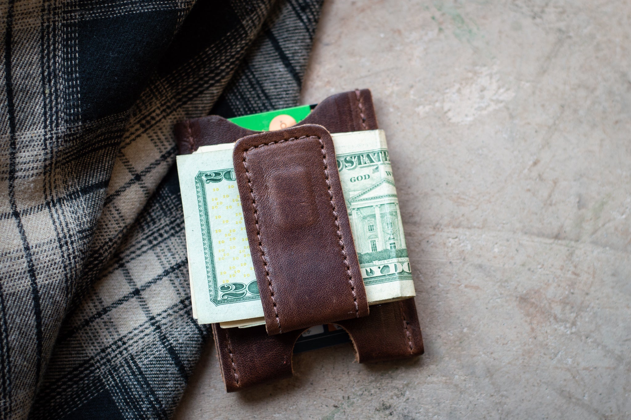 Leather money clip secures folded twenty-dollar bills and a credit card near a plaid jacket.
