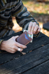 A man's hands pull cash from an Urban Southern leather cardholder wallet while sitting at a weathered wooden picnic table.
