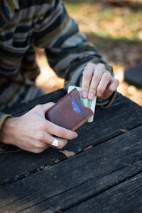 A man's hands pull cash from an Urban Southern leather cardholder wallet while sitting at a weathered wooden picnic table.
