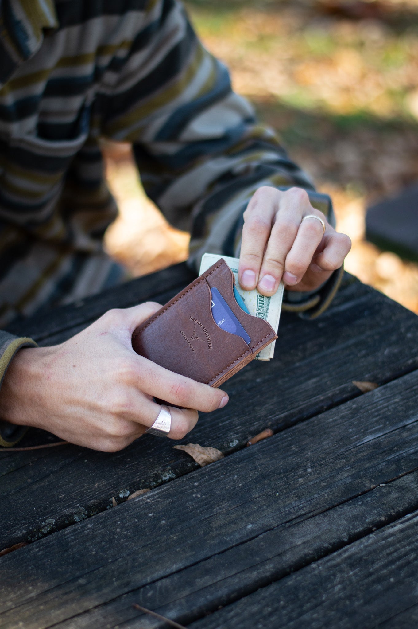 A man's hands pull cash from an Urban Southern leather cardholder wallet while sitting at a weathered wooden picnic table.
