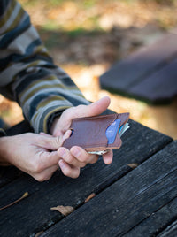 Person holds a slim brown leather wallet with debit card and cash on a weathered wooden picnic table.
