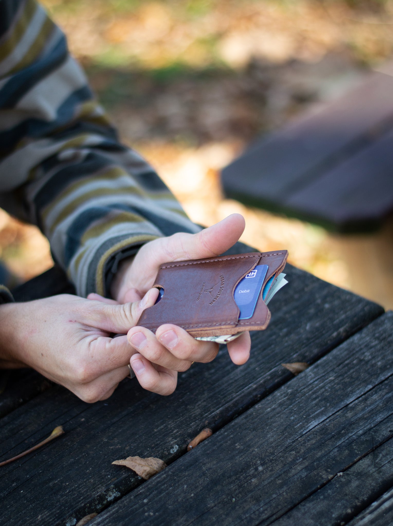 Person holds a slim brown leather wallet with debit card and cash on a weathered wooden picnic table.
