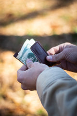 A person pulls American dollar bills from a brown leather money clip outdoors.
