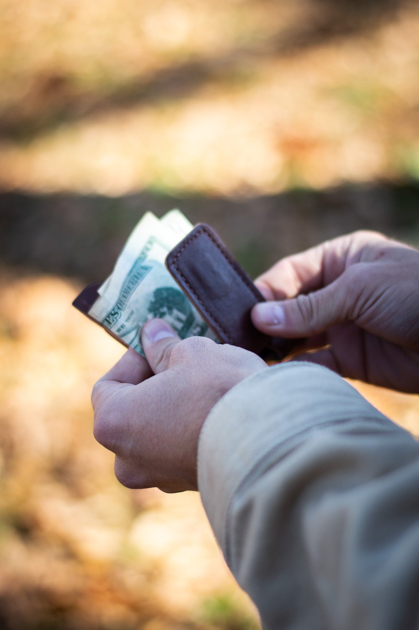 A person pulls American dollar bills from a brown leather money clip outdoors.
