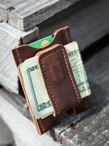 A minimalist brown leather wallet displays twenty-dollar bills and a credit card on rustic wooden planks.

