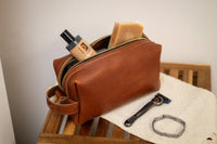 A brown leather men's dopp kit bag displays grooming essentials like cologne, soap, and a razor.
