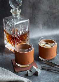 A crystal decanter sits behind two whiskey glasses, each wrapped in personalized leather sleeves, next to chilling stones and ice tongs, showcasing a refined home bar setup.
