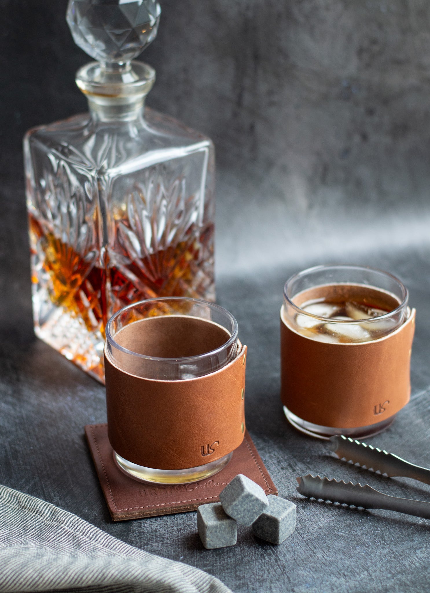 A crystal decanter sits behind two whiskey glasses, each wrapped in personalized leather sleeves, next to chilling stones and ice tongs, showcasing a refined home bar setup.
