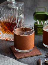 A glass of whiskey sits on a leather coaster with a matching leather sleeve, near a crystal decanter, an ammo can, and bullet-shaped accessories.
