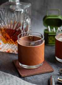A glass of whiskey sits on a leather coaster with a matching leather sleeve, near a crystal decanter, an ammo can, and bullet-shaped accessories.
