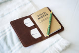 A Field Notes memo book rests inside a brown leather cover with business cards and a decorative pen on a striped cloth.
