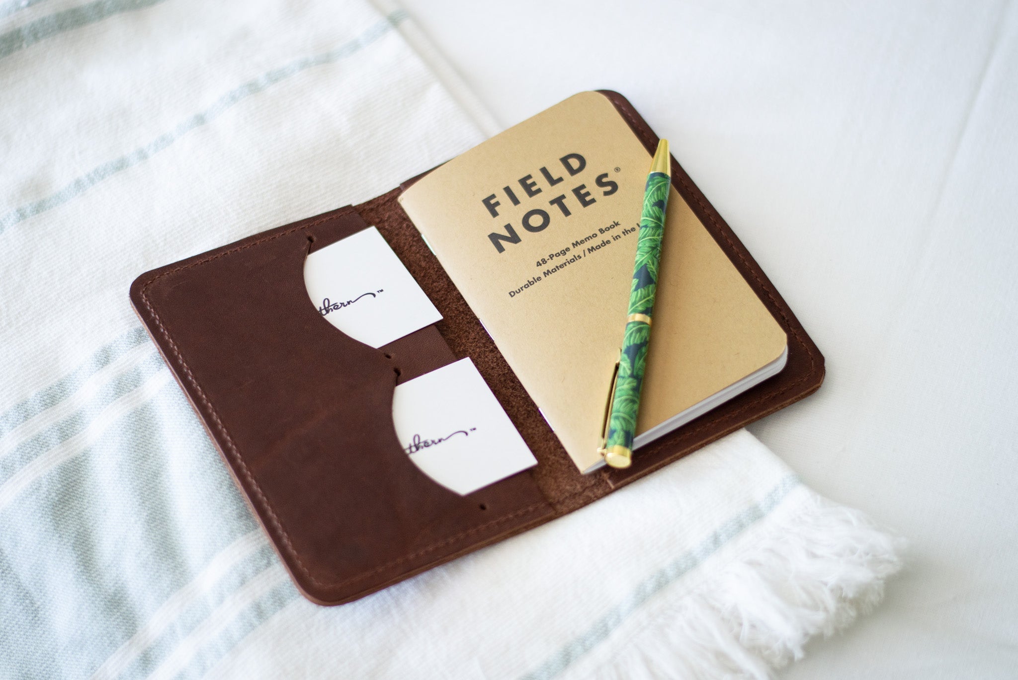 A Field Notes memo book rests inside a brown leather cover with business cards and a decorative pen on a striped cloth.