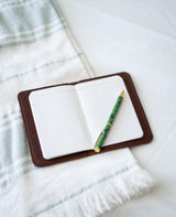 Open leather journal shows blank lined pages with a decorative green pen resting on top, set against a striped towel.
