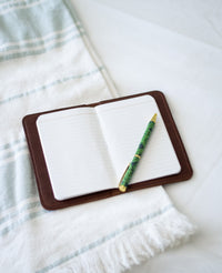 Open leather journal shows blank lined pages with a decorative green pen resting on top, set against a striped towel.
