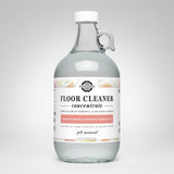A glass jug displays the label for Rustic Strength plant-based floor cleaner concentrate with lemon, lime, orange, and grapefruit essential oils.