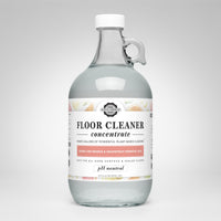 A glass jug displays the label for Rustic Strength plant-based floor cleaner concentrate with lemon, lime, orange, and grapefruit essential oils.