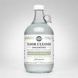 A glass jug displays Rustic Strength Floor Cleaner concentrate, highlighting its plant-based formula with rosemary, mint, and tea tree essential oils.