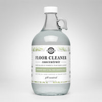 A glass jug displays Rustic Strength Floor Cleaner concentrate, highlighting its plant-based formula with rosemary, mint, and tea tree essential oils.