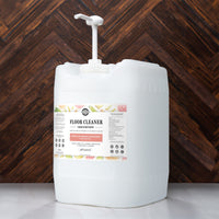 A five-gallon container of Rustic Strength floor cleaner concentrate with lemon, lime, orange, and grapefruit essential oils sits against a dark wood background.