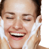 A smiling woman washes her face with foamy cleanser.
