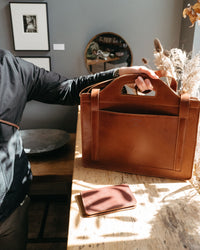 A person presents a handcrafted brown leather briefcase and matching cardholder on a weathered wooden surface, highlighting the quality craftsmanship and design.
