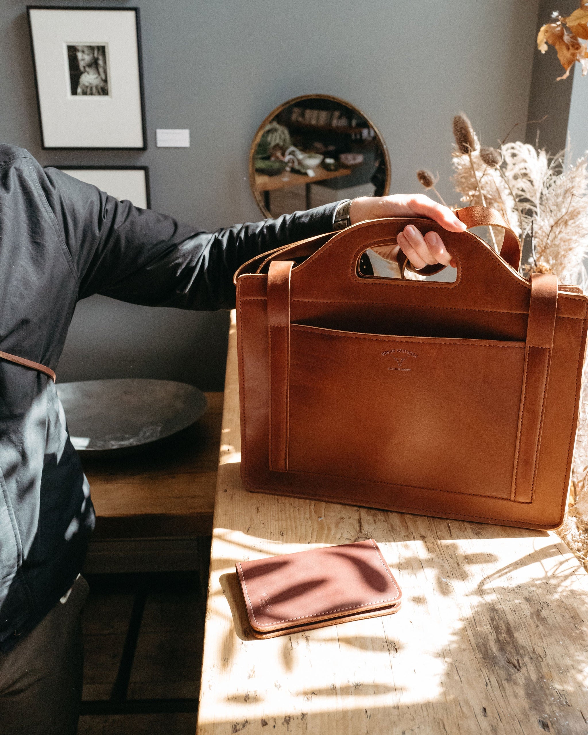 A person presents a handcrafted brown leather briefcase and matching cardholder on a weathered wooden surface, highlighting the quality craftsmanship and design.
