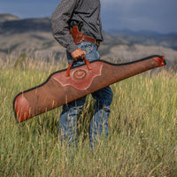Person holding a large brown leather gun case in a grassy field with mountains in the background