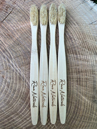 Four Raw Naturals bamboo toothbrushes lie vertically on a textured wooden surface.
