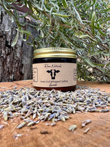 A jar of Rowe Naturals grass-fed whipped tallow lavender balm sits amidst dried lavender buds on a wooden surface with rosemary sprigs behind it.
