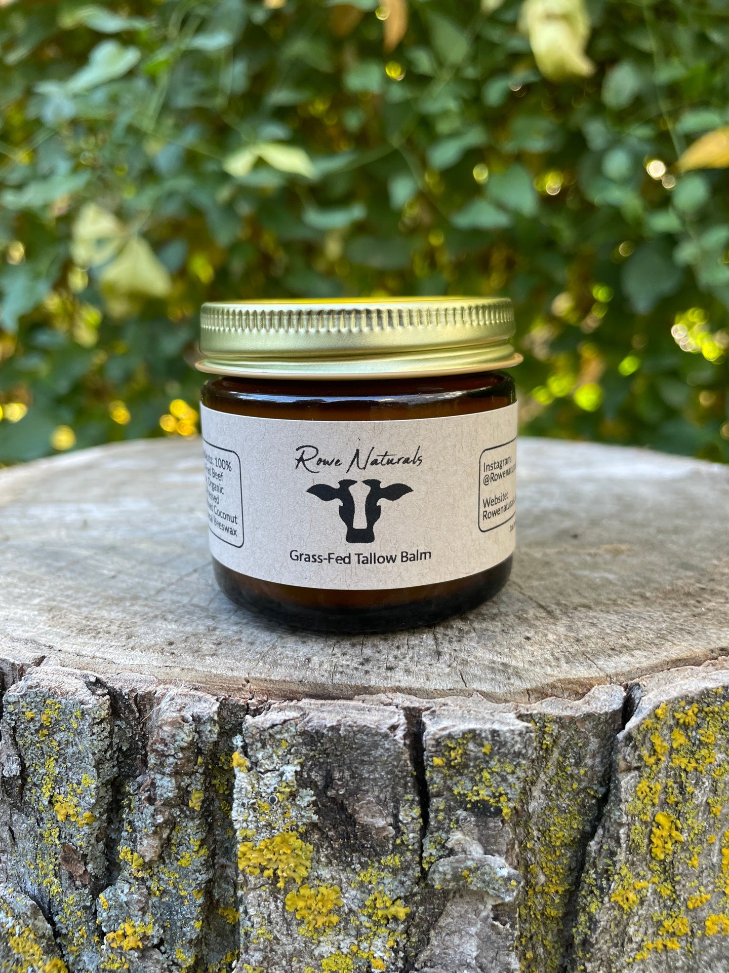 A jar of Rowe Naturals grass-fed tallow balm sits on a textured tree stump outdoors.