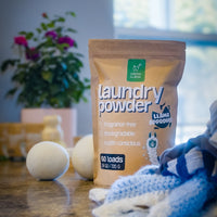 Green Llama laundry powder and wool dryer balls sit on a countertop near folded laundry.
