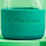Close-up shows a reusable glass bottle with a teal silicone sleeve labeled "All-Purpose" and a white straw.
