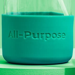 Close-up shows a reusable glass bottle with a teal silicone sleeve labeled "All-Purpose" and a white straw.
