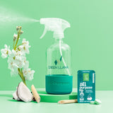 Green Llama all-purpose cleaner sprays next to a lemongrass-scented refill packet with flowers and coconut chunks arranged around the bottle.

