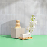 A natural cleaning still life presents a wooden dish brush atop a stone, alongside a bar of soap in a bamboo dish, accented by white flowers against a tiled backdrop.
