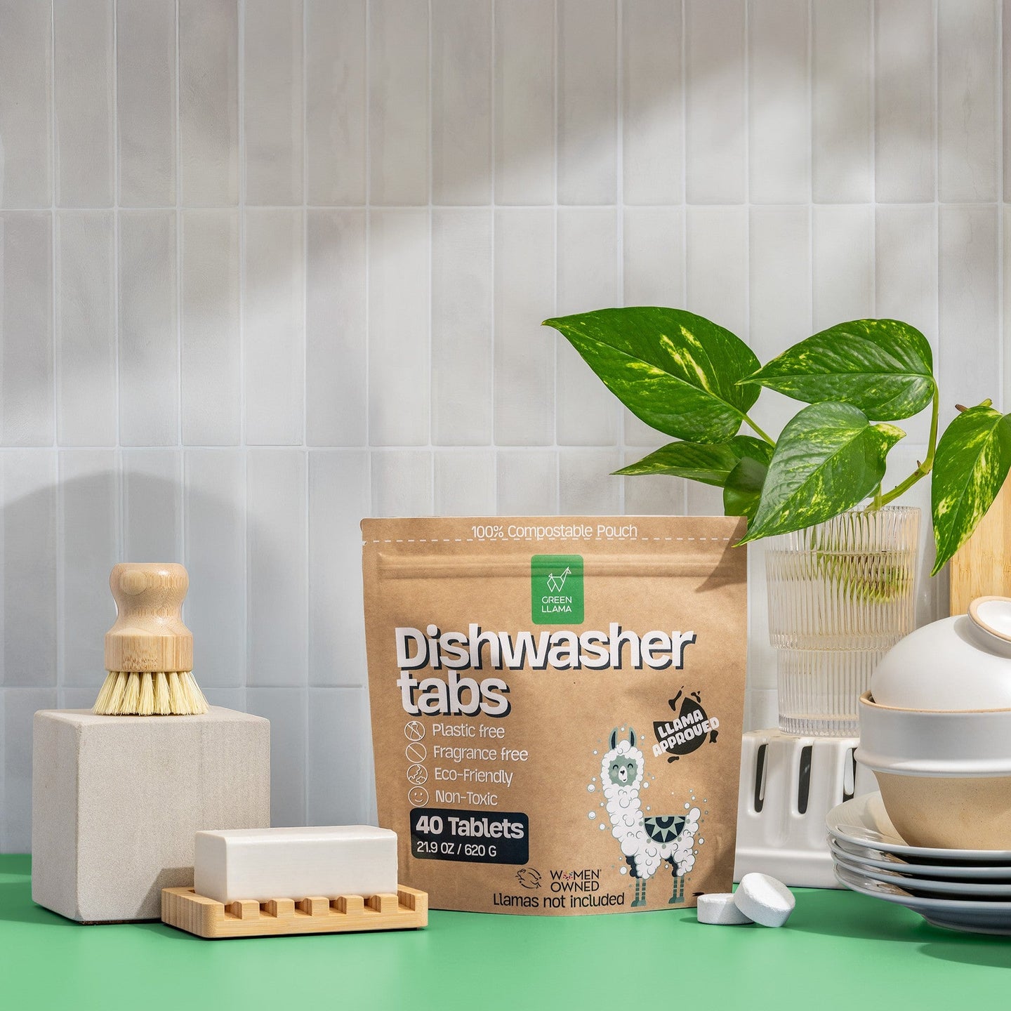 Green Llama dishwasher tabs sit on a green countertop near dishware, a cleaning brush, and a houseplant against a white tiled wall.
