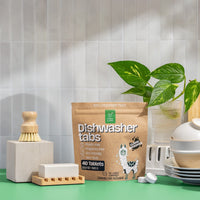Green Llama dishwasher tabs sit on a green countertop near dishware, a cleaning brush, and a houseplant against a white tiled wall.

