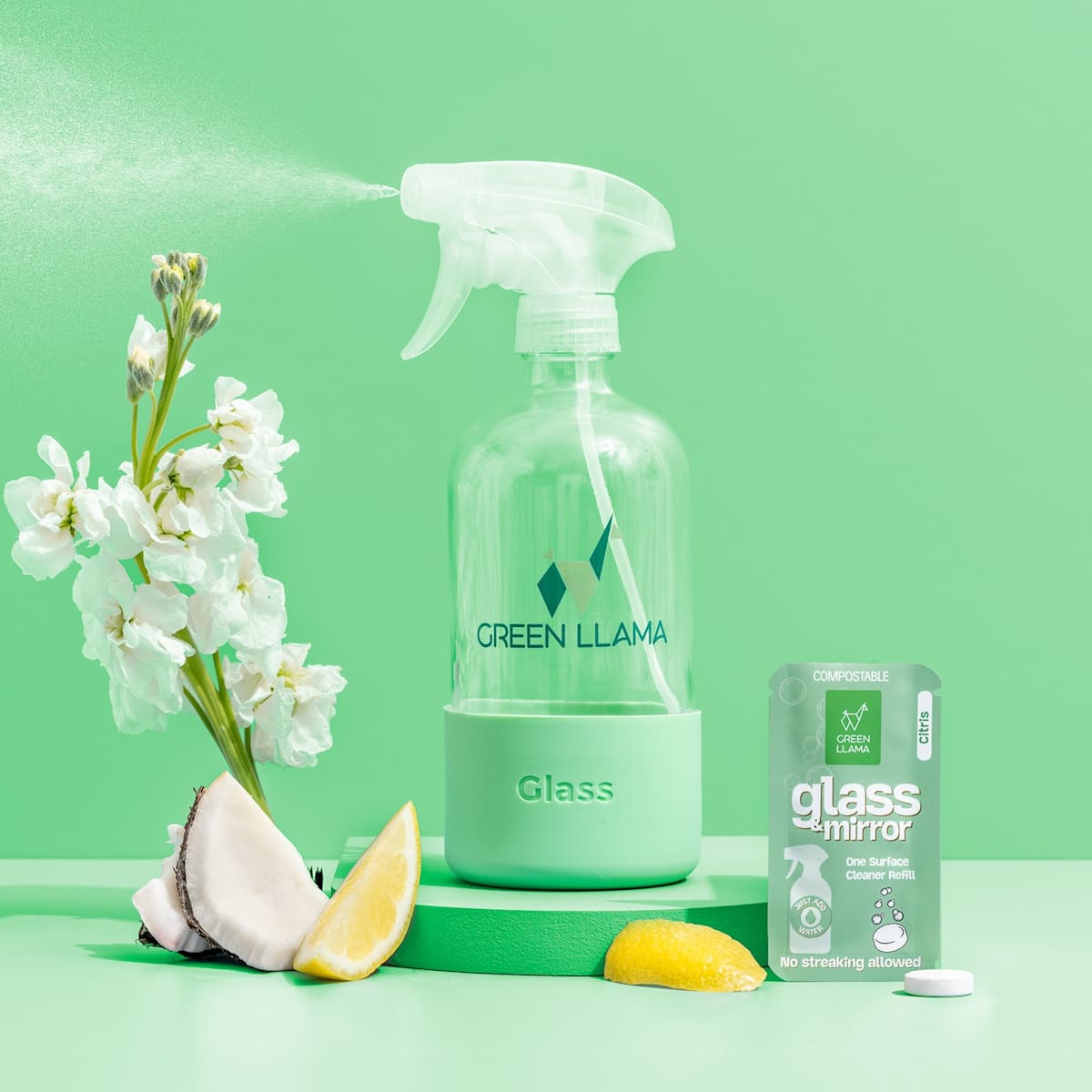 Green Llama's glass cleaner sprays beside white flowers, coconut, and lemon, showcasing their eco-friendly refill.
