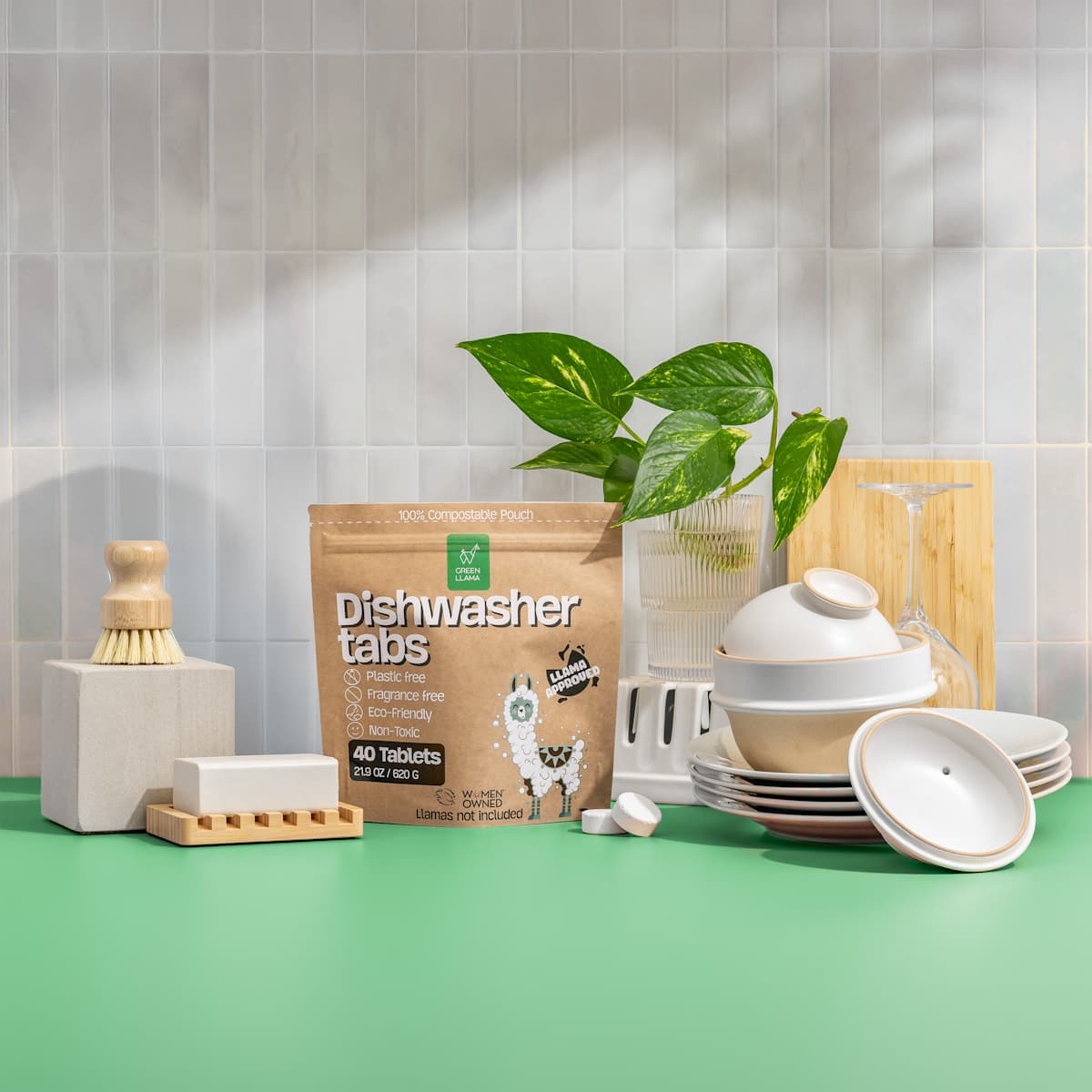 A bag of Green Llama dishwasher tabs sits among dishes, a brush, and soap on a green countertop.

