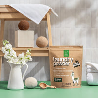 Green Llama's eco-friendly laundry powder, boasting a compostable pouch, is presented beside wool dryer balls and a white floral arrangement.
