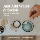 Person mixes mineral-powered oral rinse with xylitol, sea salt, and essential oils in water.
