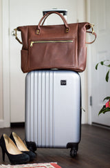 Packing stands ready with a brown leather tote bag atop a silver rolling suitcase and black high heels nearby.
