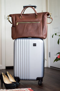 Packing stands ready with a brown leather tote bag atop a silver rolling suitcase and black high heels nearby.
