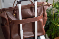 A brown leather travel bag sits on top of a rolling suitcase near a green leafy plant.
