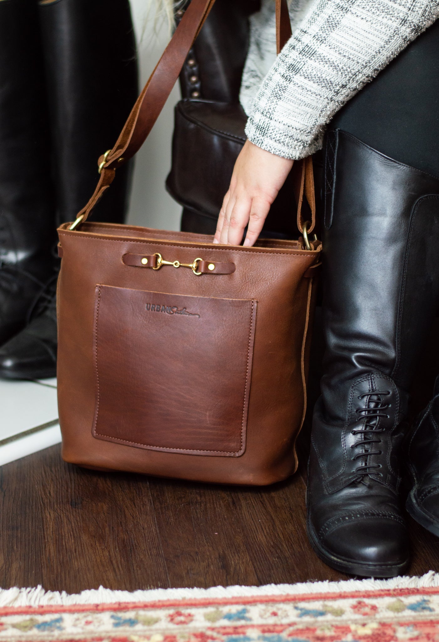 A woman reaches into her "Urban Southern" brown leather crossbody tote bag while standing near her tall, laced-up black riding boots.
