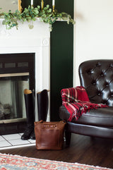 Styled equestrian-inspired leather tote bag and riding boots sit near a fireplace in a cozy, decorated room.
