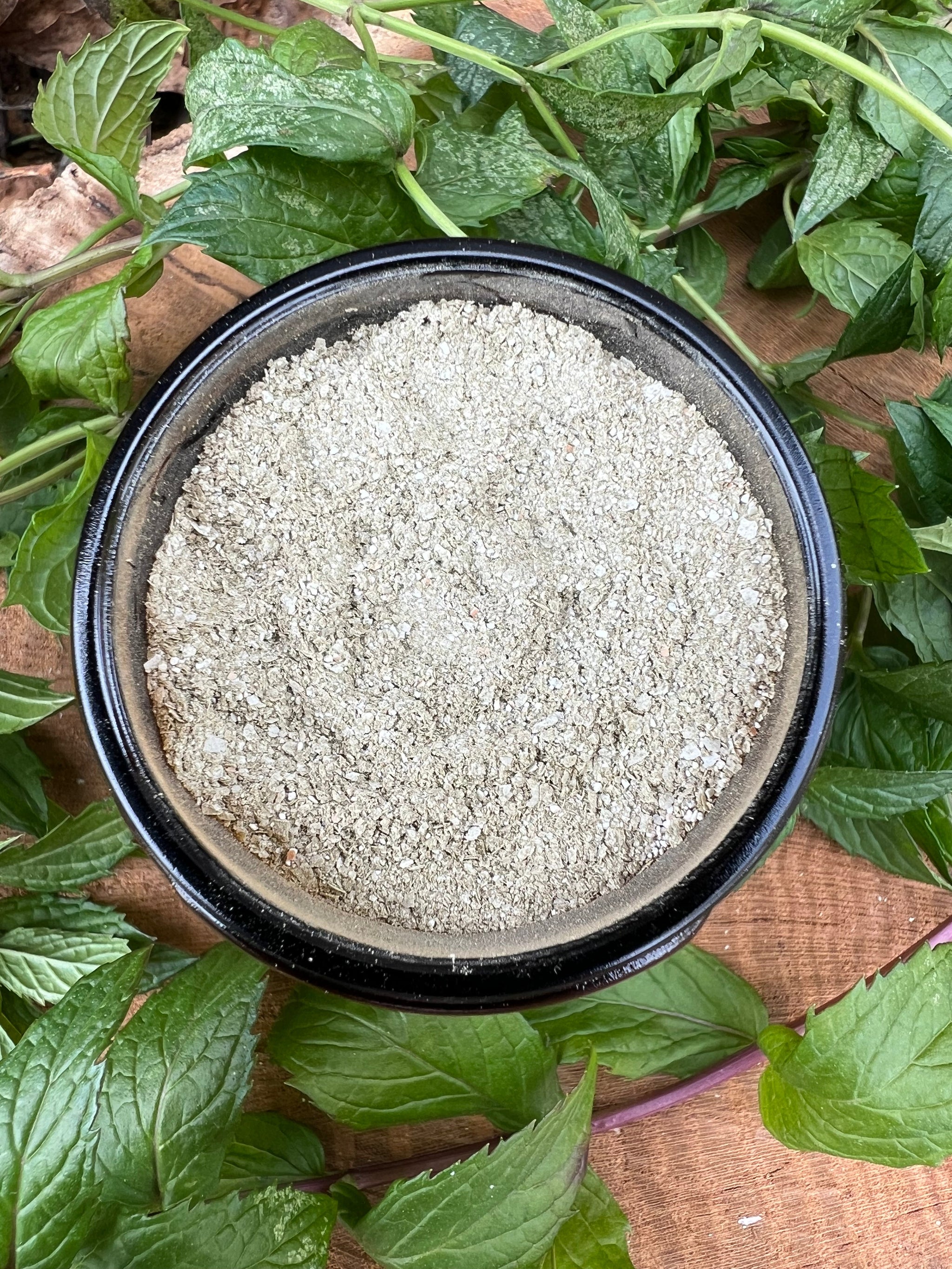 A black bowl holds crushed oyster shells fertilizer, surrounded by fresh variegated mint leaves on a wooden surface.
