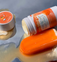 Breathe Naturals orange creamsicle deodorant and matching lip balm sit beside a melting orange popsicle on a reflective surface, creating a summer-themed beauty still life.
