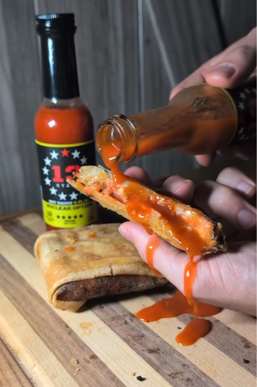 Hot sauce being poured onto a sausage on a wooden board with a bottle of hot sauce in the background.